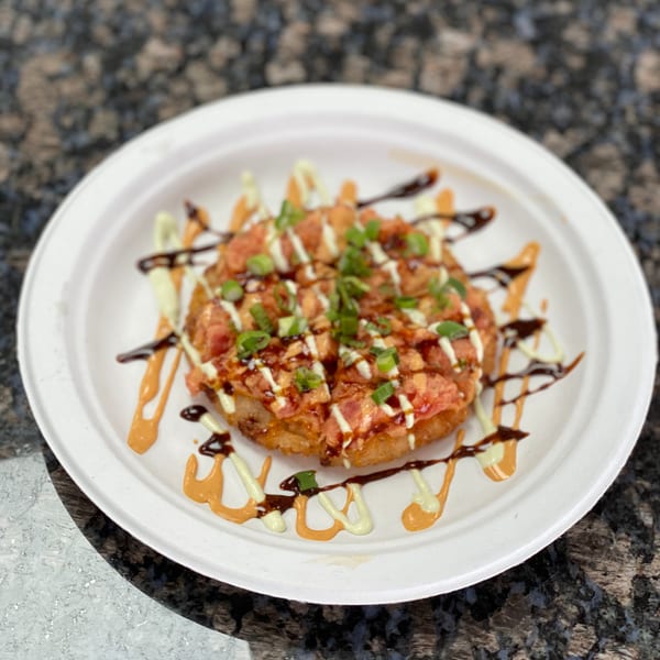 Round fried dish topped with diced pink fish, green onions, and drizzled sauces on a white plate.