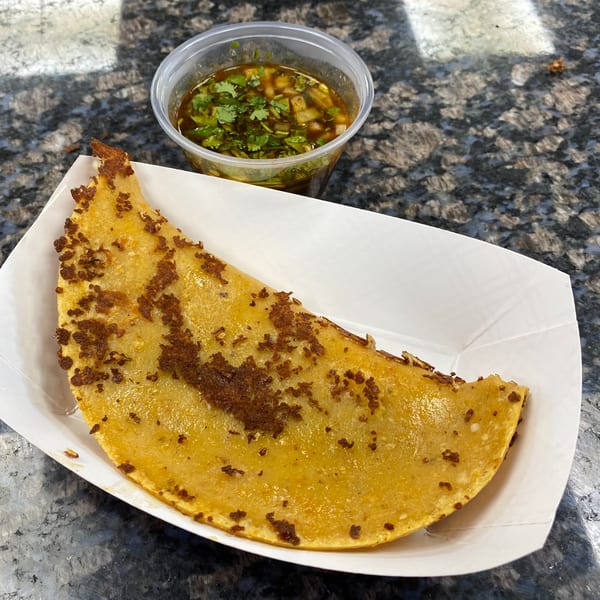 A golden-brown quesadilla with crispy bits in a white tray, next to a cup of green salsa.